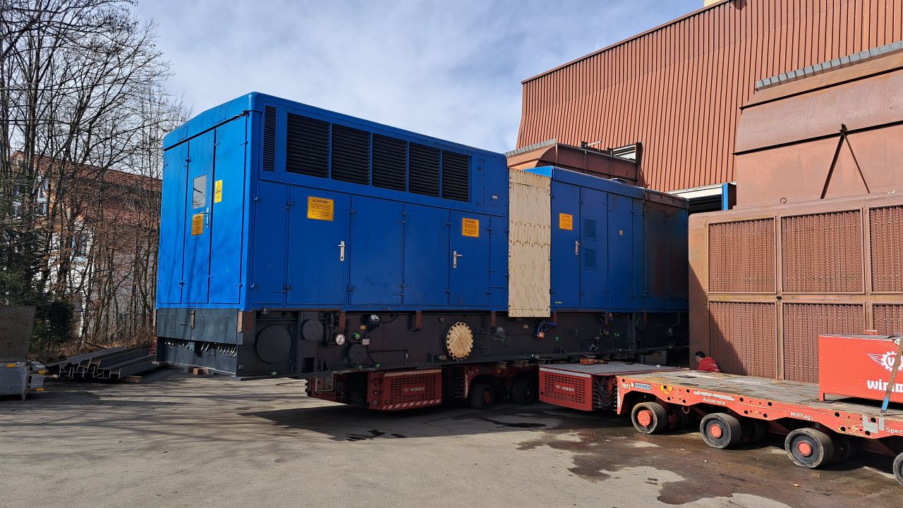 Large blue generator emerging from the Hausham power plant building onto a red multi-axle trailer.