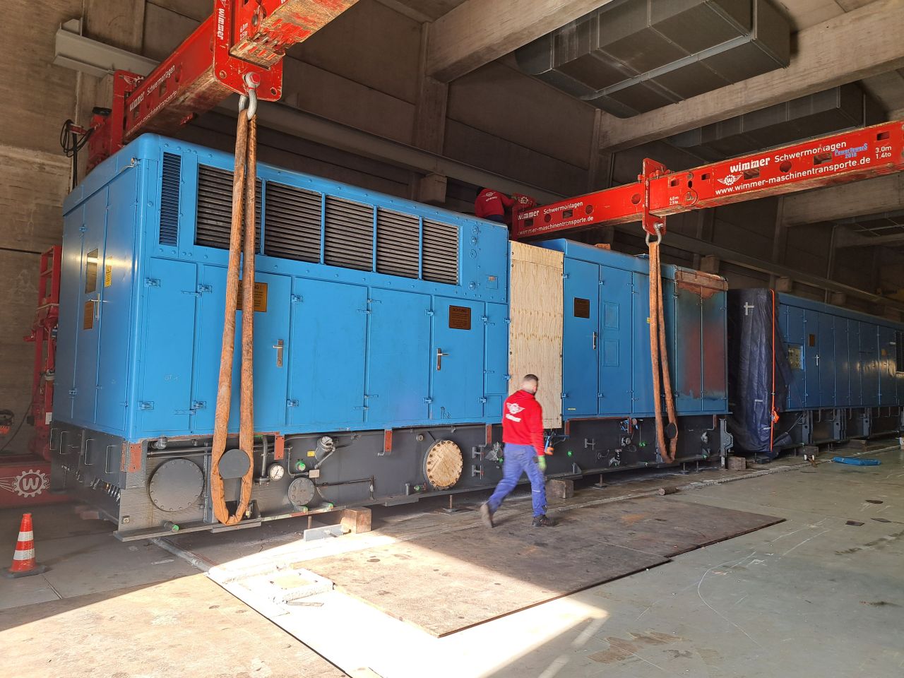 Workers using heavy lift gantry cranes and straps to secure a large blue generator inside the Hausham facility.